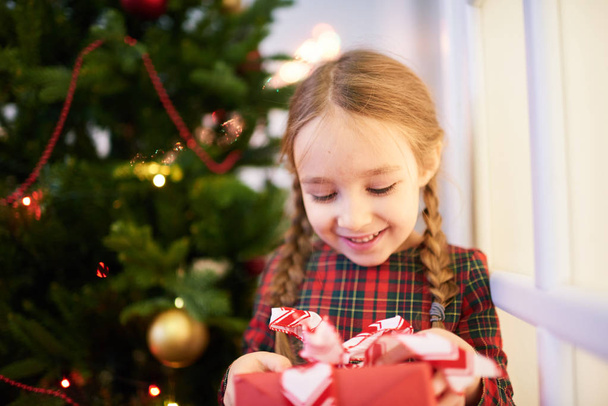 Smiling little girl wearing tartan dress opening gift box while standing at decorated Christmas tree, portrait shoot - Foto, afbeelding