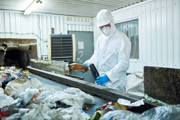 Portret van werknemer dragen van biohazard pak en veiligheidshelm werken bij afval verwerkingsbedrijf sorteren van recycleerbare materialen op transportband - Foto, afbeelding