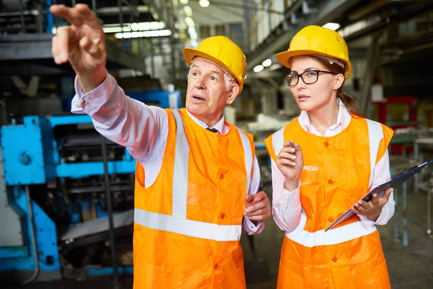 Portrait of senior factory worker explaining something to young woman pointing away at machines,  both wearing hardhats and reflective jackets - Photo, Image