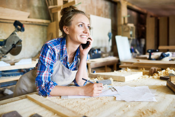 Cheerful young carpenter discussing order details with her customer on smartphone and taking necessary notes, interior of spacious workshop on background - Photo, Image