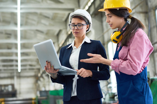 Attractive young technician wearing hardhat and overall and her middle-aged superior with laptop in hands taking inventory at production department of modern plant - Photo, Image