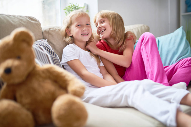Portrét dvou šťastné malé sestřičky sedět na pohodlné pohovce v útulném obývacím pokoji doma a usmívá se na kameru - Fotografie, Obrázek