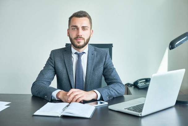 Confident bearded entrepreneur wearing classical suit looking at camera while sitting at desk, interior of modern office on background, waist-up portrait shot - Photo, Image