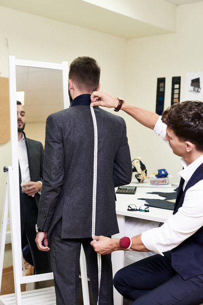 Portrait of young tailor measuring back of handsome man fitting bespoke suit in traditional atelier studio - Фото, изображение