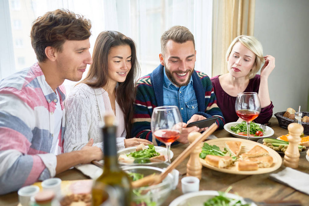 Portrait of four young people smiling happily watching videos from smartphone at dinner table during holiday celebration - Photo, Image