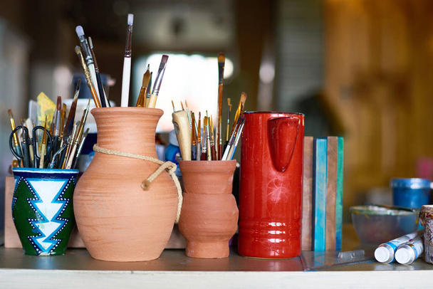 Bodegón fondo de varias ollas de arcilla con pinceles y suministros de arte en la mesa en el pequeño estudio de arte y artesanía
 - Foto, Imagen