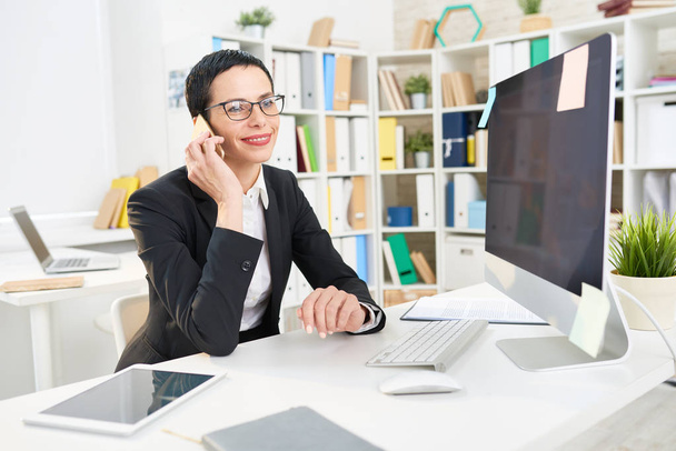 Улыбающийся предприниматель средних лет, сидящий за столом во время телефонных переговоров с деловым партнером, интерьер современного офиса на заднем плане
 - Фото, изображение
