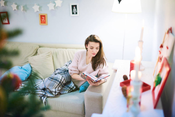 Αρκετά μελαχρινός γυναίκα κάθεται στον άνετο καναπέ και διαβάζοντας το αγαπημένο βιβλίο με ενδιαφέρον, λαμβάνοντας παράλληλα το υπόλοιπο μετά γιορτάζει τα Χριστούγεννα με τους συγγενείς - Φωτογραφία, εικόνα