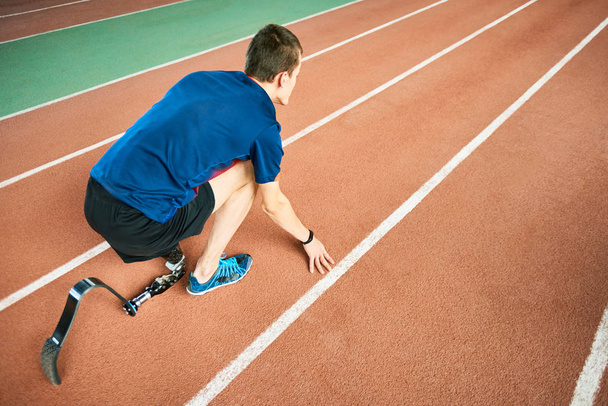 High angle portrait of young amputee athlete on start position on running track in modern indoor stadium - Photo, Image