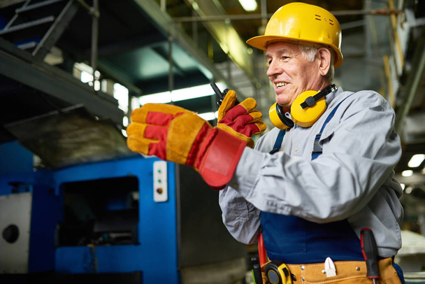 Retrato del trabajador de la fábrica senior usando un equipo de radio portátil y sonriendo hablando con alguien en el taller
 - Foto, imagen