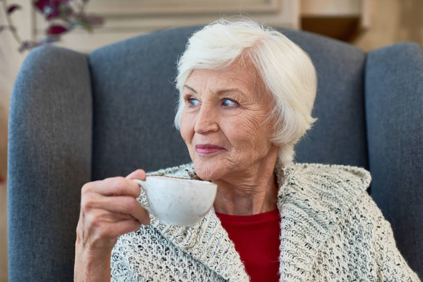 Retrato de mujer mayor elegante sonriendo mirando hacia otro lado sentado en una silla grande y cómoda con taza de té
 - Foto, Imagen