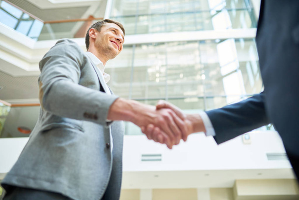 Vue à angle bas de partenaires d'affaires souriants serrant la main tout en se tenant debout dans le hall de bureau moderne spacieux
 - Photo, image