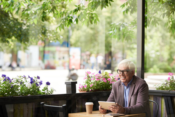 Portret van lachende senior man zit op café tafel buiten bedrijf digitale tablet, kopie ruimte - Foto, afbeelding