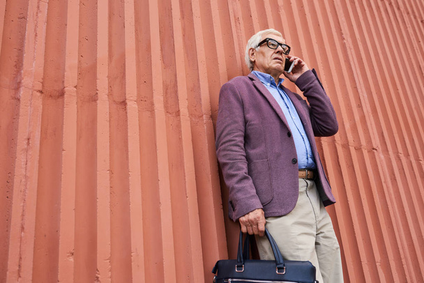 Low angle portrait of trendy senior man speaking by smartphone standing by bright orange wall outdoors - 写真・画像