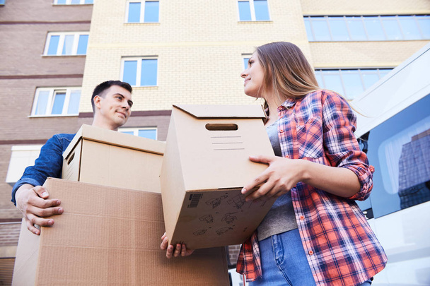 Low angle portrait of happy couple holding cardboard boxes while moving into new home, outdoors - Foto, imagen