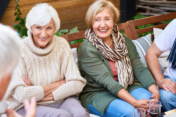hübsche Seniorinnen mit charmantem Lächeln, die ihrem männlichen Freund interessiert zuhören, während sie sich im gemütlichen kleinen Patio treffen - Foto, Bild