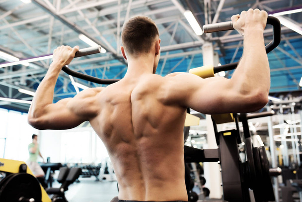 Vista posterior retrato de un joven guapo con músculos de bombeo en el pecho desnudos haciendo ejercicio en la máquina en el gimnasio
 - Foto, Imagen