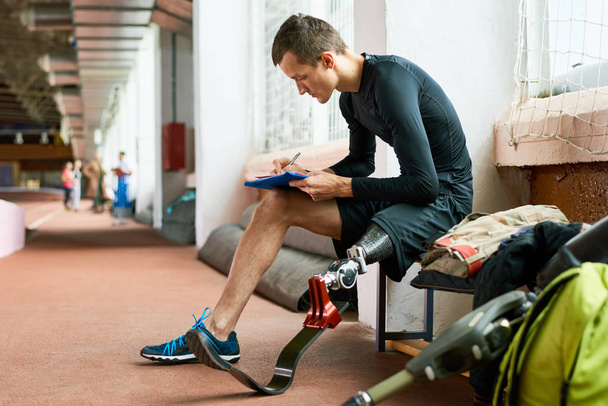 Side view portrait of young amputee athlete relaxing on bench in gym and writing something, copy space - Photo, Image