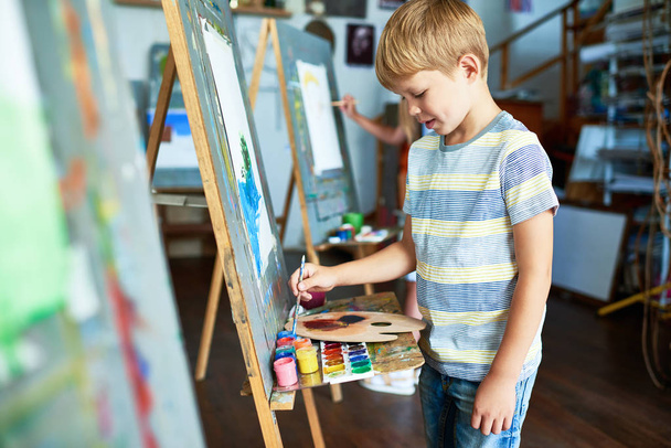 Portrait of  blonde little boy painting with watercolors on easel during art class in studio - Foto, imagen