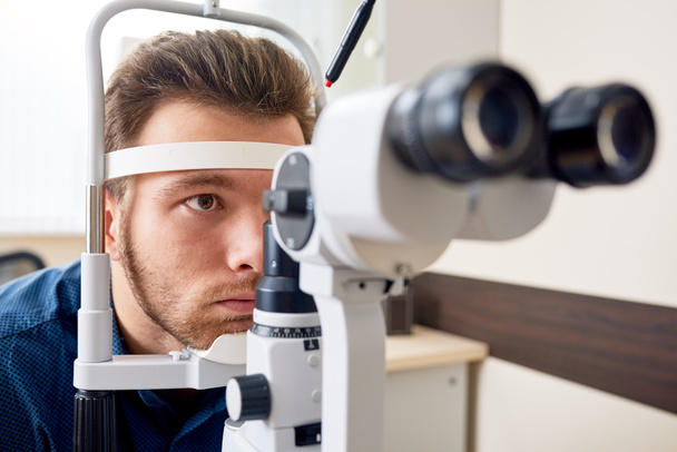 Portrait of young man  looking at slit lamp machine, resting head on stand during sight testing in optometrist office - Foto, imagen