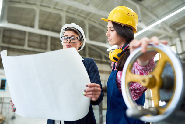 Smart female technologist reading industrial blueprint while talking to female manual worker in hardhat in factory workshop - Photo, Image