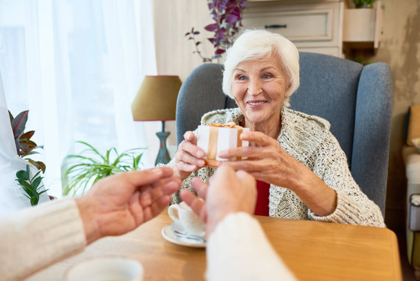 Portrait d'un heureux couple de personnes âgées échangeant un cadeau assis à table, mettant l'accent sur une heureuse femme âgée recevant une boîte-cadeau
 - Photo, image