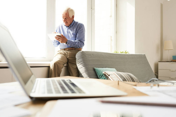 Портрет современного пожилого человека, сидящего дома на подоконнике, ноутбук на переднем плане
 - Фото, изображение