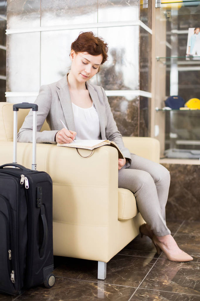Täyspitkä muotokuva tyylikäs nuori liikenainen odottaa hotellin aulassa ja tehdä työmuistiinpanoja istuu sohvalla matkalaukut
 - Valokuva, kuva