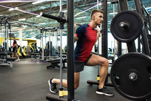 Портрет сильного молодого человека, занимающегося тяжелой атлетикой с штангой на стенде во время тренировки в современном тренажерном зале, копировальное пространство
 - Фото, изображение