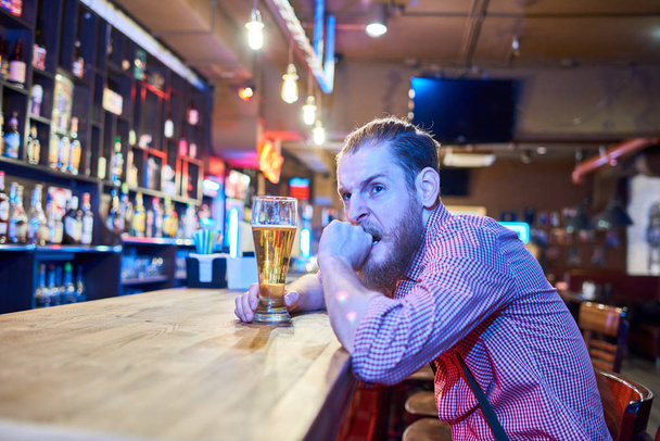 Retrato de homem barbudo ansioso olhando preocupado e irritado enquanto assiste a jogos esportivos na TV sentado no balcão do bar no bar com alto copo de cerveja, espaço de cópia
 - Foto, Imagem