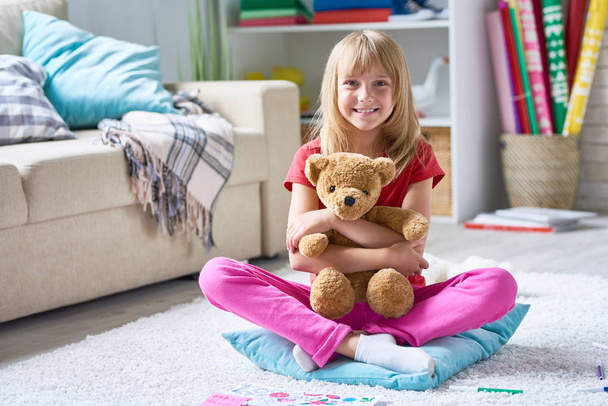 Volledige lengte portret van vrij klein meisje op zoek naar camera met toothy glimlach terwijl zittend in de lotuspositie op tapijt en knuffelen pluchen teddybeer, interieur van gezellige woonkamer op achtergrond - Foto, afbeelding