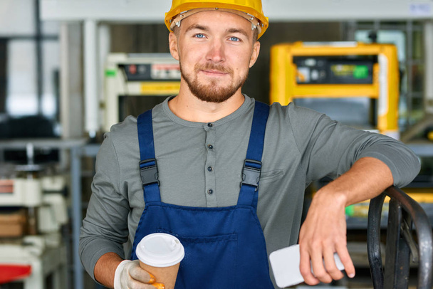 Portret młodego pracownika przerwę w warsztacie fabrycznie patrząc na kamery trzymając kubek kawy i uśmiechając się - Zdjęcie, obraz