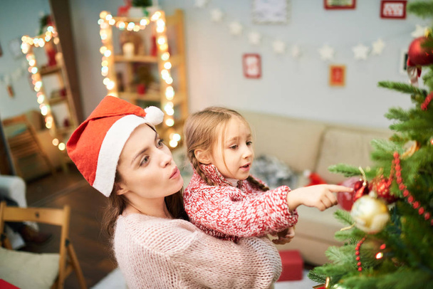 Attractive woman wearing Santa hat holding her little daughter on arms while decorating Christmas tree together, interior of living room on background - Foto, afbeelding