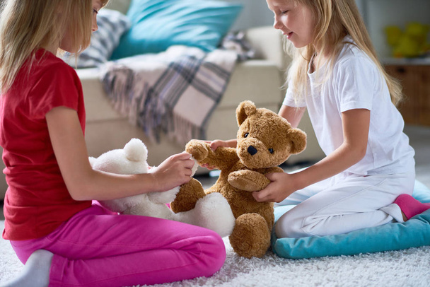 Side view of pretty little sisters playing with plush teddy bears while sitting on cozy carpet at spacious living room - Foto, Imagem
