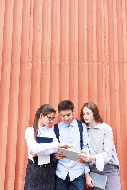Grupo de adolescentes estudantes colaborando no projeto da escola com a ajuda de tablet digital, enquanto em pé contra a parede de concreto vermelho
 - Foto, Imagem