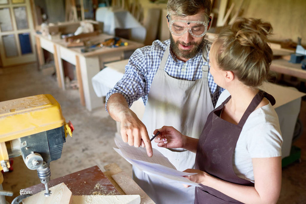 High angle portrait of bearded carpenter explaining something  to his female assistant in modern woodworking shop - Valokuva, kuva