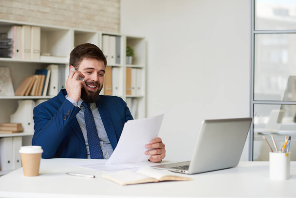 Portrét úspěšných vousatý podnikatel hovoří telefonicky a usmíval se při práci u stolu v moderní kanceláři - Fotografie, Obrázek
