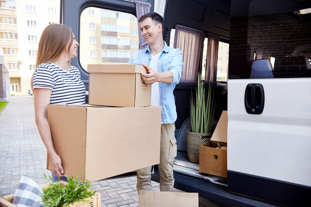 Portrait of smiling young man handing cardboard box to wife unloading moving van outdoors - Photo, Image