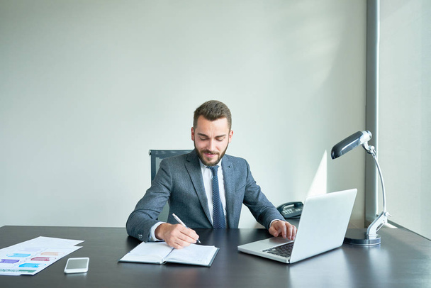 Mladý vousatý podnikatel v klasického obleku zapisování poznámek, zatímco sedí v kanceláři a analýzou finančních údajů s pomocí notebooku, pas-stránky na výšku - Fotografie, Obrázek