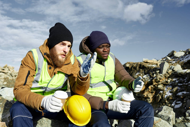 Portrait von zwei Industriearbeitern in reflektierenden Jacken, einer davon afrikanisch, die es sich in der Kaffeepause bei der Arbeit gemütlich machen und auf der Minenbaustelle im Freien plaudern - Foto, Bild