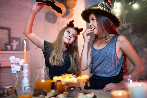 Portret van twee tienermeisjes dragen kostuums van Halloween, spelen met speelgoed vleermuis uit ingerichte kamer tijdens feest - Foto, afbeelding