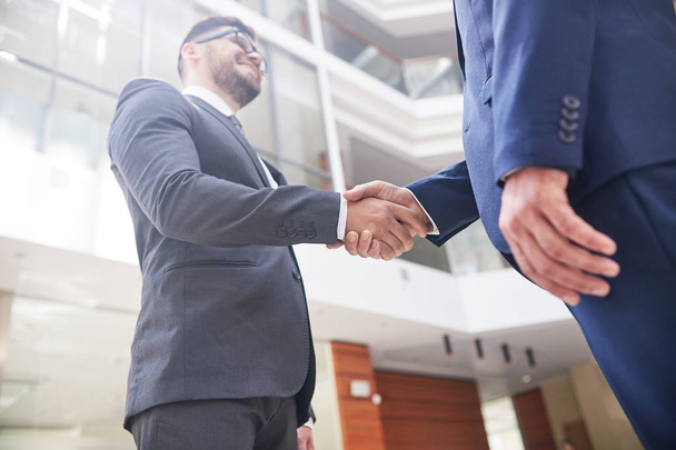 Confident business partners shaking hands after successful completion of negotiations, interior of modern office lobby on background, low angle view - Photo, Image