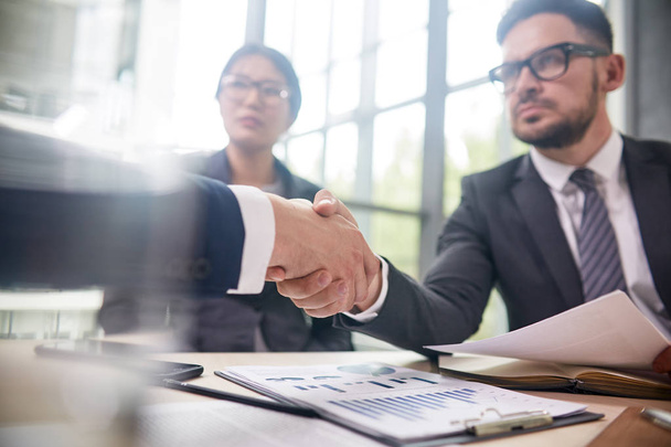 Confident bearded entrepreneur wearing elegant suit sitting at boardroom table and greeting business partner with firm handshake, pretty Asian assistant manager next to him - Photo, Image
