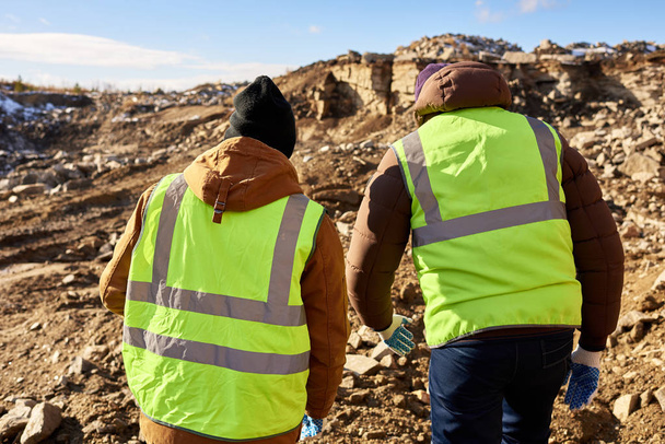 Back view portrait of two industrial workers wearing reflective jackets, one of them African, inspecting mineral mines on worksite outdoors - Photo, Image