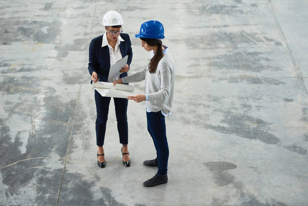 Confident businesswoman wearing hardhat studying blueprint with help of pretty young industrial engineer while carrying out inspection at production department of modern plant - Photo, Image