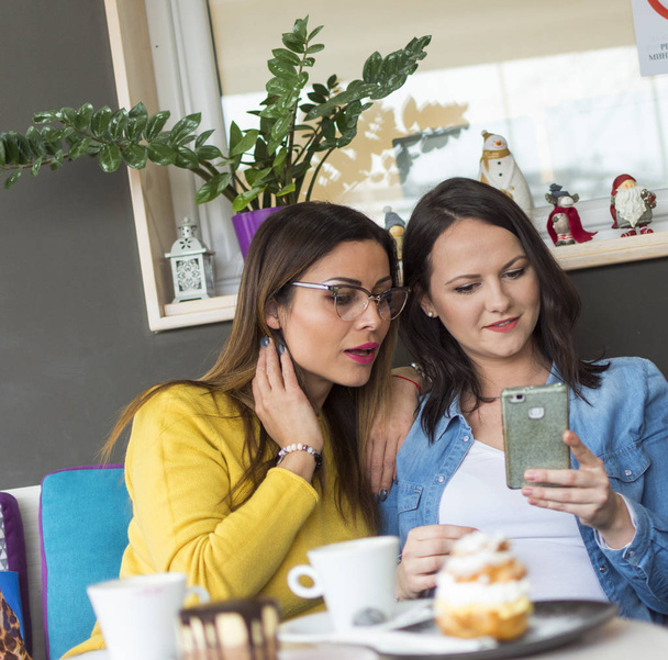 portrét dvou dospělých dcer / dvě dospělé usmívající se dívky jíst koláče a sleduje události na sociálních sítích, na mobilní telefon - Fotografie, Obrázek