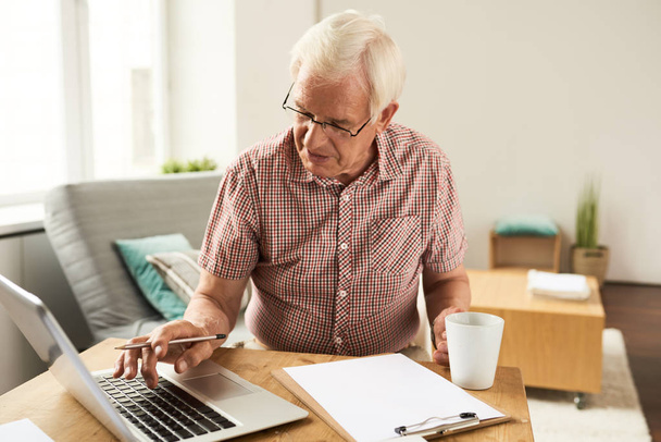 Портрет пожилого человека, работающего с ноутбуком дома, подсчитывающего финансы и печатающего
 - Фото, изображение