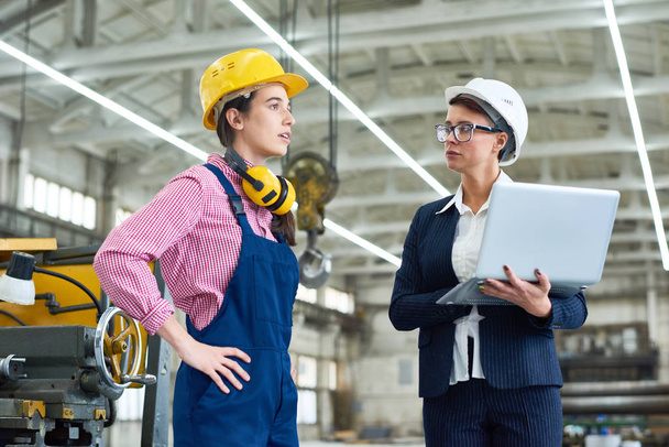 Serious confident female engineer in formal suit talking to manual worker with protective ear muffs on neck working in factory workshop - Photo, Image