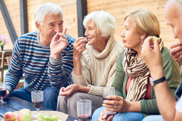 Grupo de amigos mayores que pasan un rato agradable juntos en la fiesta del patio trasero: charlan animadamente entre sí, beben vino tinto y comen frutas frescas
 - Foto, Imagen