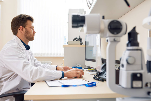 Side view portrait of  male ophthalmologist  using PC sitting at desk in office and typing on keyboard, optometric equipment in foreground - Фото, изображение